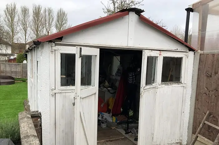 A weathered white timber garage with peeling paint, a red apex roof and open doors, full of stored belongings, in a back garden awaiting removal